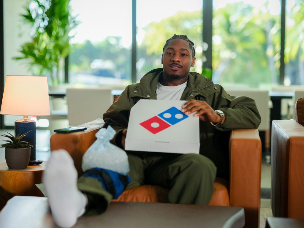 Image of Stefon Diggs on couch with ice on knee with Dominos box on lap. for Franchise News about emergency pizza