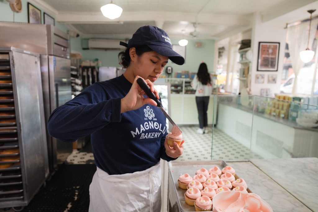 Magnolia Bakery team member icing a cupcake with the signature swirl. Vanilla cupcake pink and peach toned icing in a bakery with team member wearing dark blue branded hat and shirt.