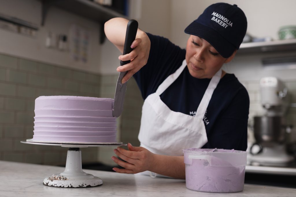 Magnolia Bakery team member Icing a Cake with purple icing and a tool.