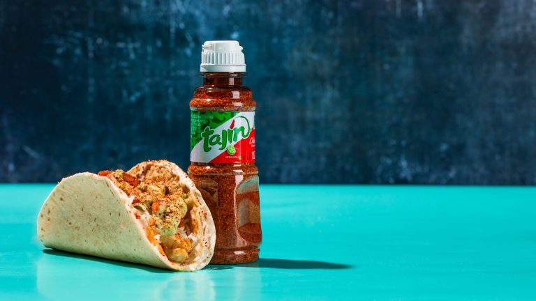 Queso Crunch Taco filled with Tequila Lime Chicken and topped with Tajín beside a Tajín bottle on a teal surface.