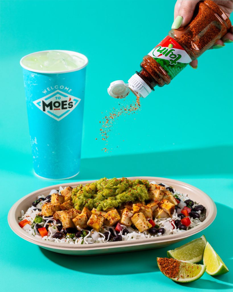 Hand sprinkling Tajín Chile Lime seasoning over a Moe’s bowl with Tequila Lime Chicken, guacamole, rice, beans, and lime wedges.